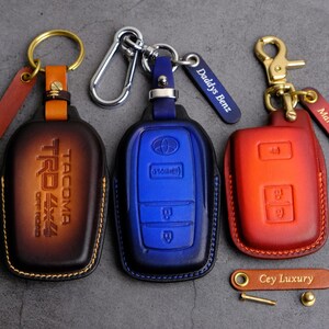 May include: Three leather key covers with different colors and designs. The first is brown with the text "TACOMA TRD OFF ROAD" in white. The second is blue with the text "TOYOTA" in white. The third is red with the text "Marron" in white. The covers are attached to keychains with different styles.