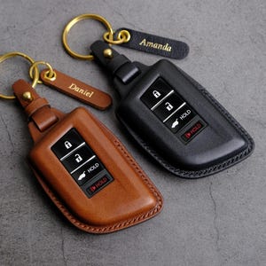 May include: Two leather key fobs with key rings and personalized name tags. The key fobs are brown and black and have a design that resembles a car key. The name tags read "Daniel" and "Amanda".