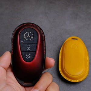 May include: A red leather key fob with a silver Mercedes-Benz logo and three buttons. The key fob is in a hand, and a yellow leather key fob is in the background.
