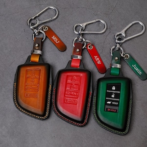 May include: Three leather car key fob covers in orange, red, and green with silver metal hardware and carabiner clips. Each cover features embossed button icons and a matching leather tag with engraved text reading MGJ, ARW, and Justin.