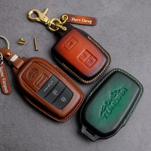 May include: Three leather keychains with personalized names. The keychains are brown, green, and orange. The keychains have the words "Harry Dawg", "Jaime Vasquez", and "TUNDRA" embossed on them.