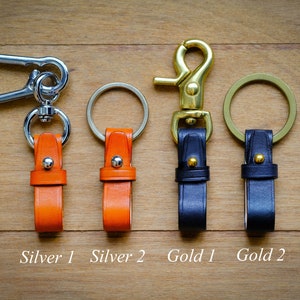 May include: Four leather keychains with metal rings and hooks. Two keychains are orange leather with silver hardware, and two keychains are black leather with gold hardware. The keychains are labeled "Silver 1", "Silver 2", "Gold 1", and "Gold 2".