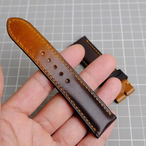 May include: A brown leather watch strap with a tan leather edge and yellow stitching. The strap is shown on a hand.
