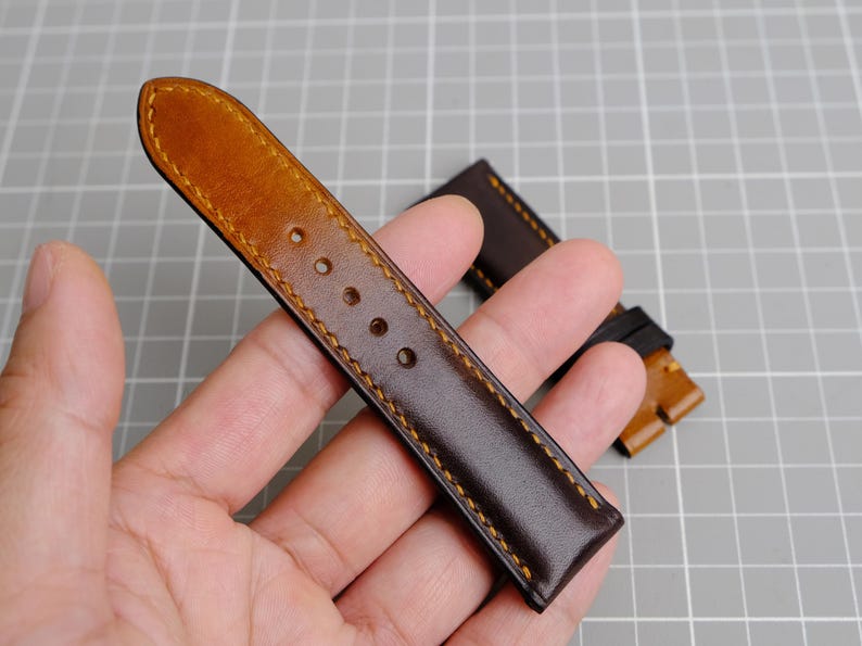 May include: A brown leather watch strap with a tan leather edge and yellow stitching. The strap is shown on a hand.