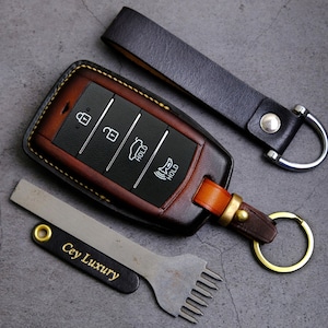 May include: A brown and black leather car key fob cover with key ring and a matching leather strap. The key fob has black buttons with white icons. A metal tool and a leather tag with the text "Cey Luxury" are also in the image.
