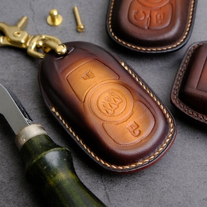 Może przedstawiać: Zbliżenie brązowego skórzanego breloka z ozdobnymi złotymi szwami. Brelok ma przyczepiony łańcuszek do kluczy i posiada wypukły wzór trzech trójkątów w kółku.