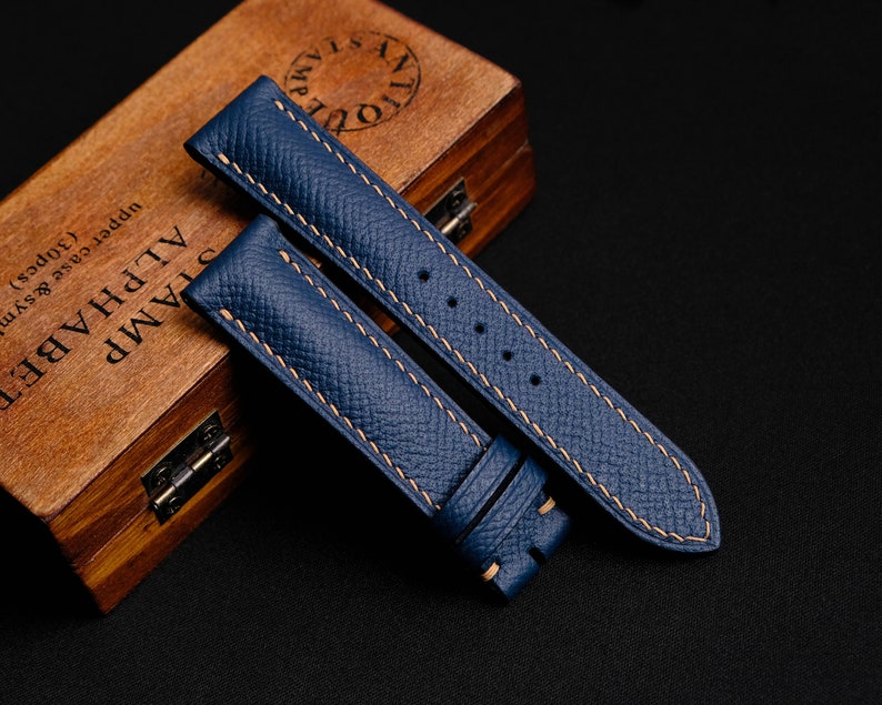 May include: Two blue leather watch straps with contrasting light brown stitching. The straps are laid out on a black surface.