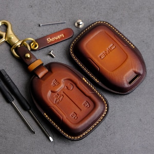 以下が含まれることがあります： 縁にステッチが施された、茶色の革製のキーホルダーが2つ。1つのキーホルダーには「Showers」という文字が刻印されています。もう1つのキーホルダーには「GMC」という文字が刻印されています。