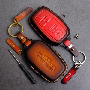 May include: Two leather key fob covers, one red and one brown, with keychains and a screwdriver. The red cover has a Toyota logo and button icons. The brown cover is embossed with "Tacoma". Accessories include keychains.