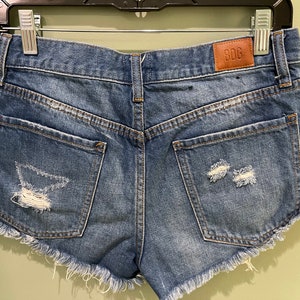 K&ouml;nnte beinhalten: Blaue Jeans-Shorts mit ausgefransten Kanten und Destroyed-Details. Die Shorts haben zwei Ges&auml;&szlig;taschen und ein braunes Leder-Patch mit dem Text "BDC" auf der rechten Ges&auml;&szlig;tasche.