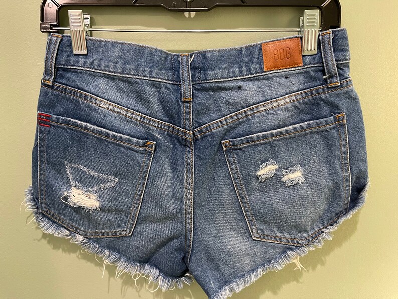 K&ouml;nnte beinhalten: Blaue Jeans-Shorts mit ausgefransten Kanten und Destroyed-Details. Die Shorts haben zwei Ges&auml;&szlig;taschen und ein braunes Leder-Patch mit dem Text "BDC" auf der rechten Ges&auml;&szlig;tasche.