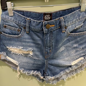 K&ouml;nnte beinhalten: Zerrissene Jeans-Shorts mit ausgefransten Kanten und einem BDG-Logo-Etikett. Die Shorts sind in einem hellen Blauton mit sichtbaren Rissen und L&ouml;chern.