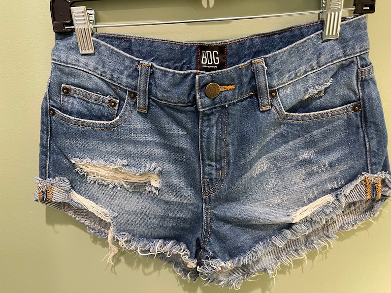 K&ouml;nnte beinhalten: Zerrissene Jeans-Shorts mit ausgefransten Kanten und einem BDG-Logo-Etikett. Die Shorts sind in einem hellen Blauton mit sichtbaren Rissen und L&ouml;chern.