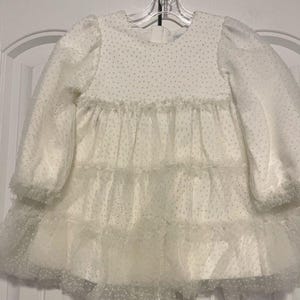 May include: A white baby dress with long, puffy sleeves and a tiered skirt. The dress features a polka dot pattern on the bodice and skirt, with delicate ruffles and a tulle overlay. The dress is hanging on a clear hanger.