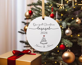 Pode incluir: Ornamento de Natal redondo branco com texto preto que diz "Our first Christmas Engaged 2024" com uma árvore de Natal estilizada em preto e branco com um coração vermelho no topo. O ornamento está pendurado em uma árvore de Natal.