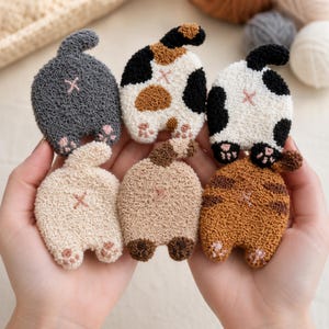 May include: Six punch needle cat butt patches featuring various fur patterns including solid gray, calico, black and white spotted, cream, light brown, and orange tabby. Each patch displays a raised tail, small pink paw pads, and an embroidered X.