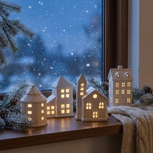 May include: A set of four white ceramic house-shaped tea light holders with cut-out windows and doors, illuminated from within, arranged on a wooden windowsill alongside frosted pine branches and a textured beige knit fabric.