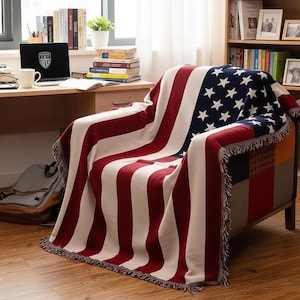 Op de afbeelding: Een decoratieve deken met het ontwerp van de Amerikaanse vlag, gedrapeerd over een fauteuil. De deken heeft rode en witte strepen en een blauwe hoek met witte sterren. De fauteuil heeft een patchwork ontwerp aan de zijkant.