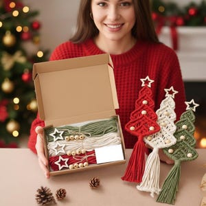 Könnte beinhalten: Ein Bastelset mit roten, weißen und grünen Makramee-Weihnachtsbaum-Ornamenten. Das Set enthält Kordel, Holzperlen und sternförmige Holzzuschnitte. Drei fertige Bäume werden gezeigt, jeder mit Sternaufsatz und Quaste.