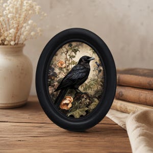 May include: An oval-shaped black frame containing a detailed illustration of a black crow perched on a branch. The bird is surrounded by muted green foliage and pale orange flowers, creating a natural scene within the dark, smooth border.