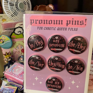 May include: A set of eight pronoun pins with black and pink designs. The pins feature different pronoun combinations, including "he/him", "she/her", "they/them", "he/they", "she/they", and "any pronouns". One pin says "ask my pronouns".