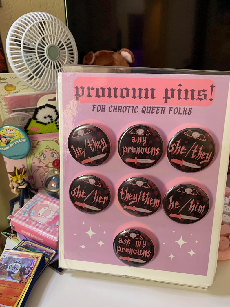 May include: A set of eight pronoun pins with black and pink designs. The pins feature different pronoun combinations, including "he/him", "she/her", "they/them", "he/they", "she/they", and "any pronouns". One pin says "ask my pronouns".