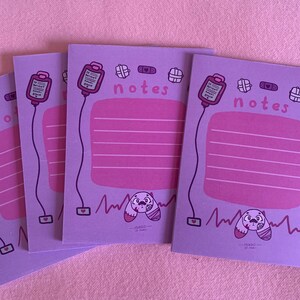 Puede incluir: Tres blocs de notas morados con una ilustraci&oacute;n rosa y blanca de un vendaje, un coraz&oacute;n, un frasco de pastillas y un perro de dibujos animados con un vendaje en la cabeza. Los blocs de notas tienen la palabra "notes" impresa en ellos.