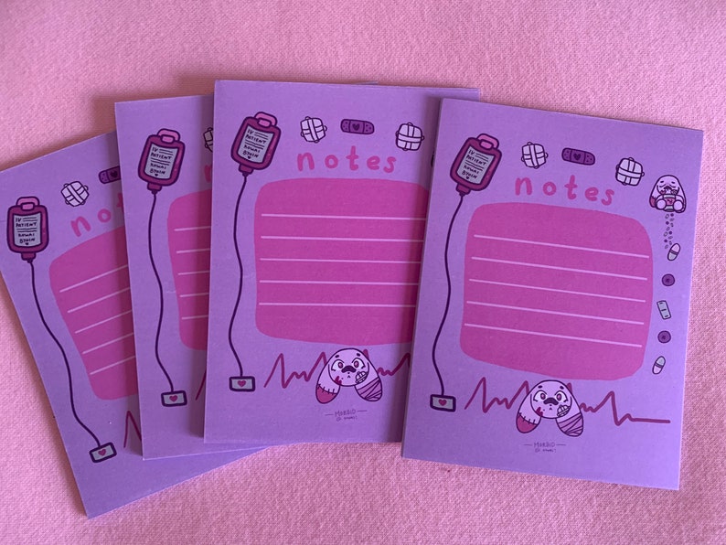 Puede incluir: Tres blocs de notas morados con una ilustraci&oacute;n rosa y blanca de un vendaje, un coraz&oacute;n, un frasco de pastillas y un perro de dibujos animados con un vendaje en la cabeza. Los blocs de notas tienen la palabra "notes" impresa en ellos.