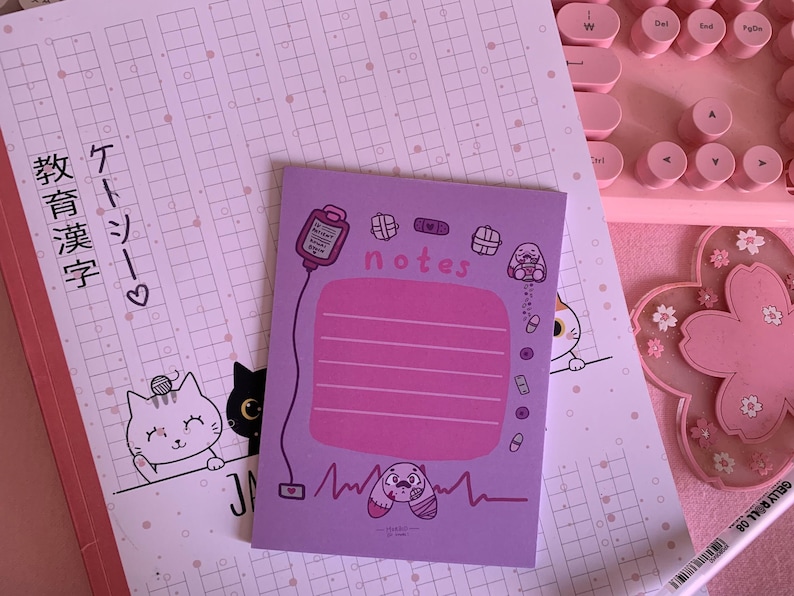 Puede incluir: Un bloc de notas morado con una secci&oacute;n de l&iacute;neas rosas para notas. El bloc de notas tiene una linda ilustraci&oacute;n de dibujos animados de un vendaje, un coraz&oacute;n y un gato con un estetoscopio. El texto "notes" est&aacute; escrito en letras blancas en el bloc de notas.