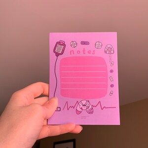 Puede incluir: Un bloc de notas rosa con una ilustraci&oacute;n de dibujos animados rosa y morado de un vendaje, un coraz&oacute;n, un frasco de p&iacute;ldoras y un conejito vendado. El bloc de notas tiene una secci&oacute;n rosa con l&iacute;neas y la palabra "notes" escrita en morado.