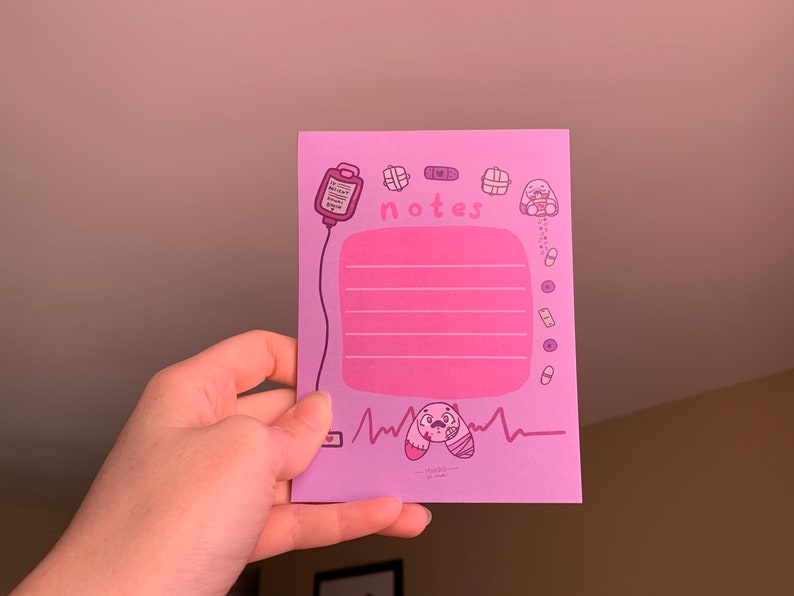 Puede incluir: Un bloc de notas rosa con una ilustraci&oacute;n de dibujos animados rosa y morado de un vendaje, un coraz&oacute;n, un frasco de p&iacute;ldoras y un conejito vendado. El bloc de notas tiene una secci&oacute;n rosa con l&iacute;neas y la palabra "notes" escrita en morado.