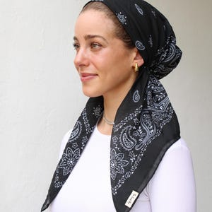 May include: Black bandana headscarf with white paisley and floral patterns. The head covering is tied at the back and worn over the head. The bandana has a small tag on the corner.