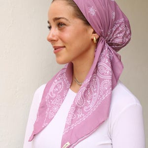 May include: A mauve-colored headscarf with a white paisley pattern. The scarf is tied at the back of the head, with the ends draped over the shoulders. The scarf is made of a lightweight fabric.