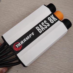 May include: A white amplifier with black text that reads "TARAMPS BASS 8K". The amplifier has a red and white logo with the word "TARAMPS" on it. The amplifier has multiple black wires coming out of it.