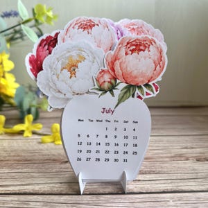May include: A desktop calendar for the month of July featuring a die-cut floral arrangement of white, pink, and deep red peony illustrations. The calendar base is white with black text displaying the days of the week and dates from 1 to 31.
