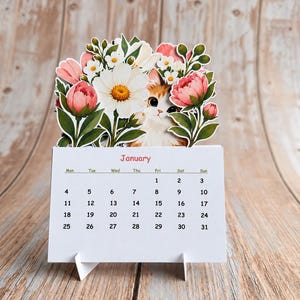 Puede incluir: Un calendario de escritorio para enero con base blanca, texto negro y un recorte decorativo. El recorte muestra la cara de un gato blanco y naranja asomándose entre peonías rosas, margaritas blancas y hojas verdes.
