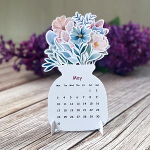 May include: A die-cut paper calendar for the month of May shaped like a jar filled with a bouquet of illustrated flowers in shades of blue, pink, and white. The calendar displays the days of the week and dates from 1 to 31 in black text.