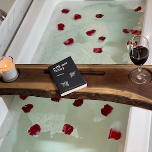 Könnte beinhalten: Ein Holz-Badetablett mit einem Glas Rotwein, einer Kerze und einem Buch mit dem Titel "milk and honey" von Rupi Kaur. Das Tablett liegt am Rand einer weißen Badewanne, die mit Wasser und Rosenblättern gefüllt ist.