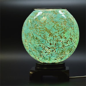 May include: A teal and gold mosaic glass globe lamp. The spherical lamp sits on a black wooden base. The glass has a cracked, textured appearance, and the lamp emits a soft glow. The lamp is plugged in with a white cord.