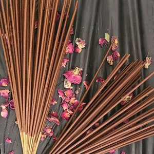 May include: Bundles of brown incense sticks are arranged on a dark surface, with dried pink rose petals scattered around them. The incense sticks are slender and have a uniform color, suggesting a natural fragrance.