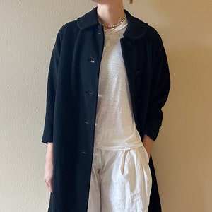 May include: A black, knee-length coat with a rounded collar and button-down closure. The coat has three-quarter sleeves and is worn over a white t-shirt and white pants. The coat appears to be made of a soft, textured fabric.