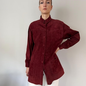 May include: A burgundy suede shirt with a button-down front, long sleeves, and two front pockets. The shirt has a collared neckline and a tailored fit. The fabric appears soft and has a slight sheen. The shirt is worn over white pants.