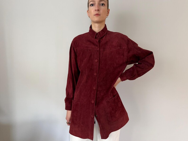 May include: A burgundy suede shirt with a button-down front, long sleeves, and two front pockets. The shirt has a collared neckline and a tailored fit. The fabric appears soft and has a slight sheen. The shirt is worn over white pants.