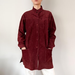 May include: A burgundy suede jacket with a mandarin collar and button-down front. The long-sleeved jacket has two large front pockets and a relaxed fit. The jacket is knee-length and appears to be vintage.