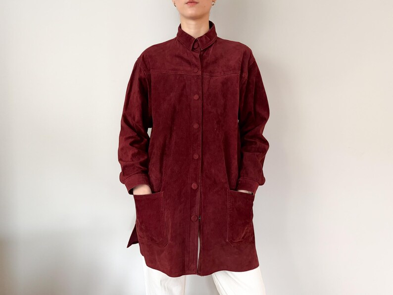 May include: A burgundy suede jacket with a mandarin collar and button-down front. The long-sleeved jacket has two large front pockets and a relaxed fit. The jacket is knee-length and appears to be vintage.