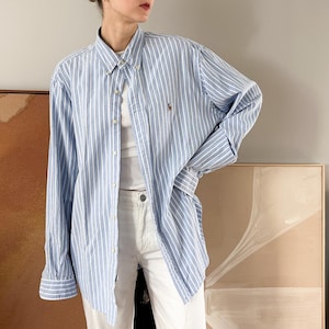 May include: A light blue and white striped button-down shirt with long sleeves. The shirt has a small logo on the chest. The shirt is worn open over a white top and paired with white pants. The shirt is a classic style.
