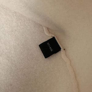 May include: Close-up of a cream-colored textile with a black tag. The tag reads "100% Wool" in white text. The fabric appears soft and textured.