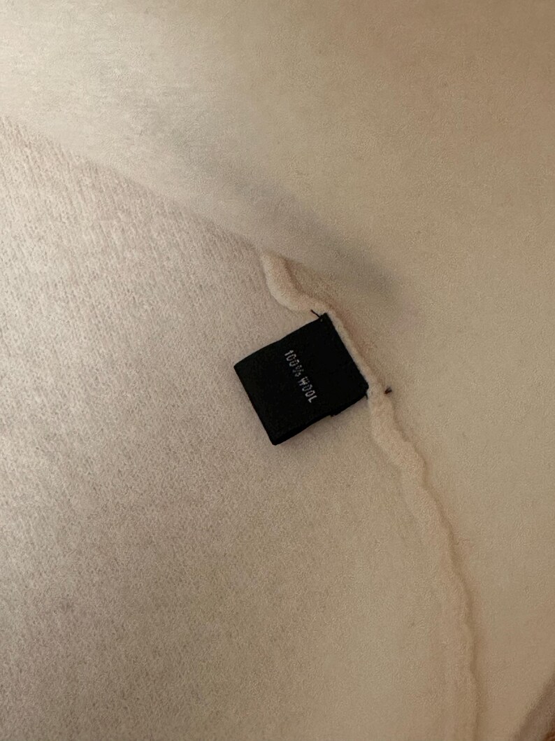 May include: Close-up of a cream-colored textile with a black tag. The tag reads "100% Wool" in white text. The fabric appears soft and textured.