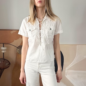 Puede incluir: Una camisa de algodón blanco con un frente de encaje y mangas cortas. La camisa tiene dos bolsillos de solapa en el pecho.