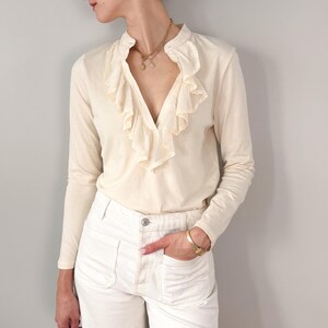 May include: A long-sleeved cream-colored shirt featuring a deep V-neckline adorned with cascading ruffles along the front placket and a small stand-up collar, paired with white denim pants that have two large front patch pockets and a button closure.